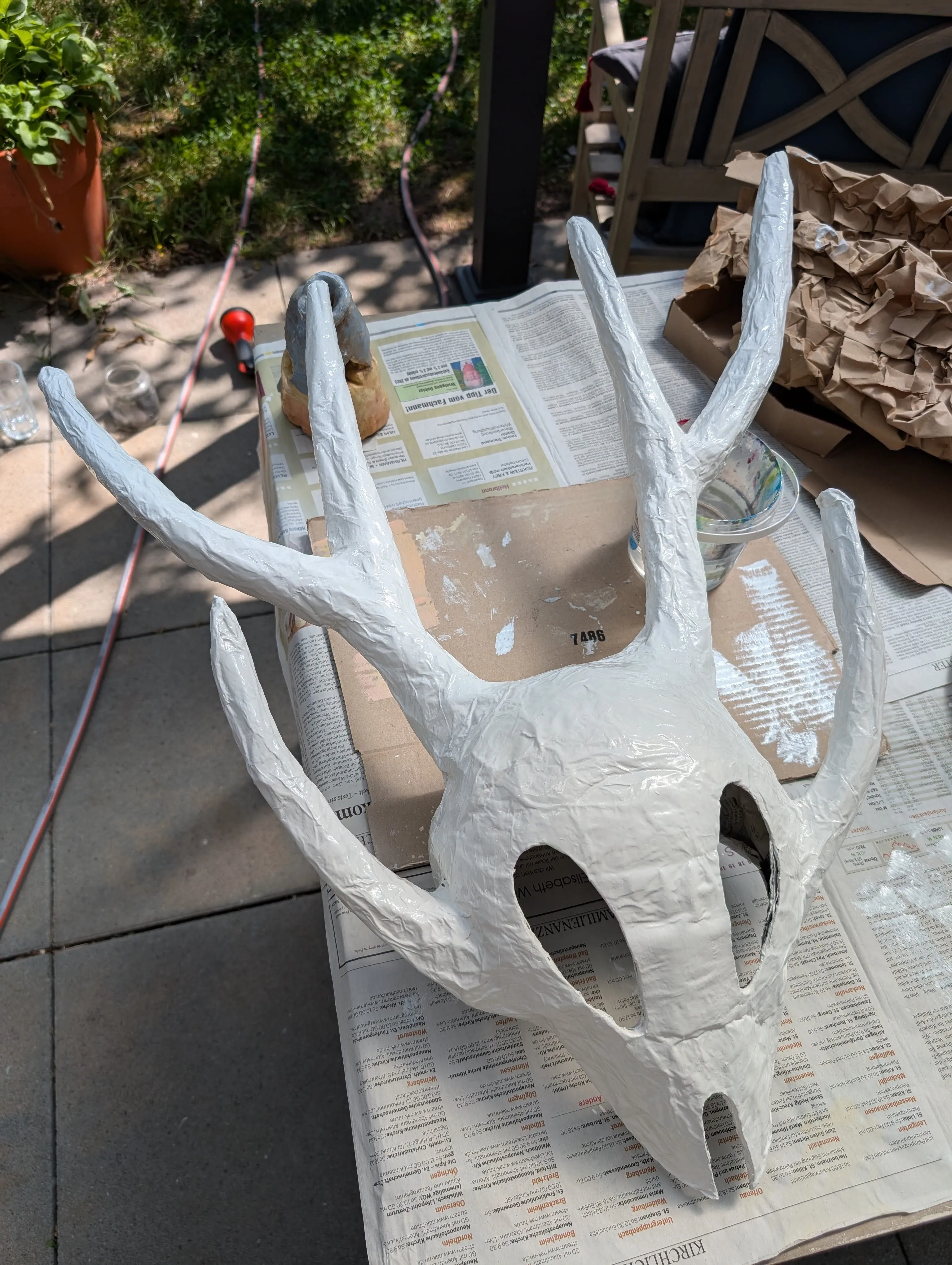 The mask made of paper mache laying on the table outside, with a layer of white paint applied.